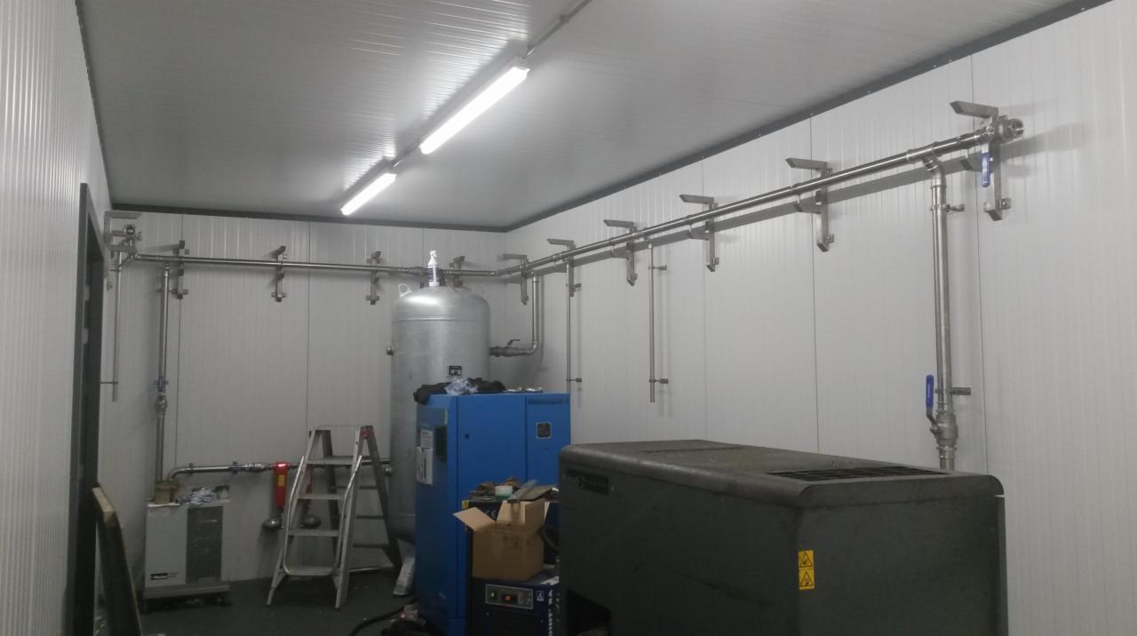Wnętrze pomieszczenia z zainstalowaną, skomplikowaną siecią rur ze stali nierdzewnej, zbiornikiem ciśnieniowym i sprężarkami, oświetlone lampami jarzeniowymi.