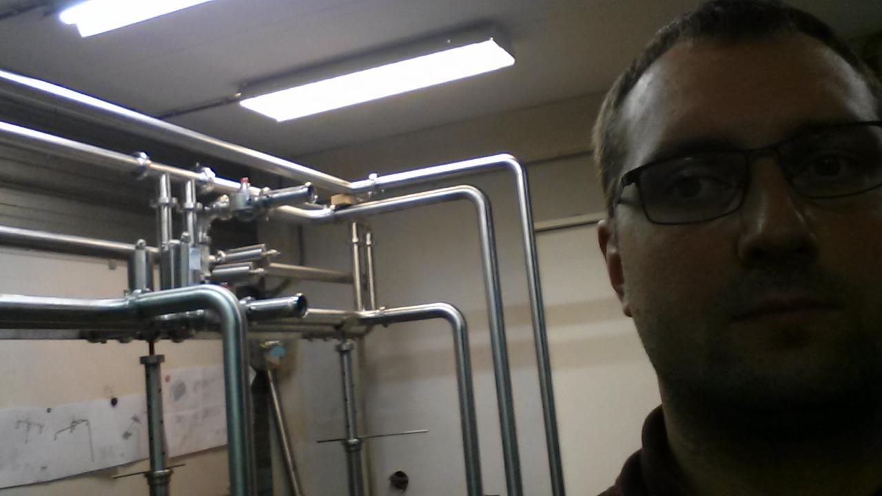 Skupisko rur ze stali nierdzewnej, prawdopodobnie instalacja przemysłowa, z fragmentem twarzy mężczyzny w okularach w prawym kadrze.