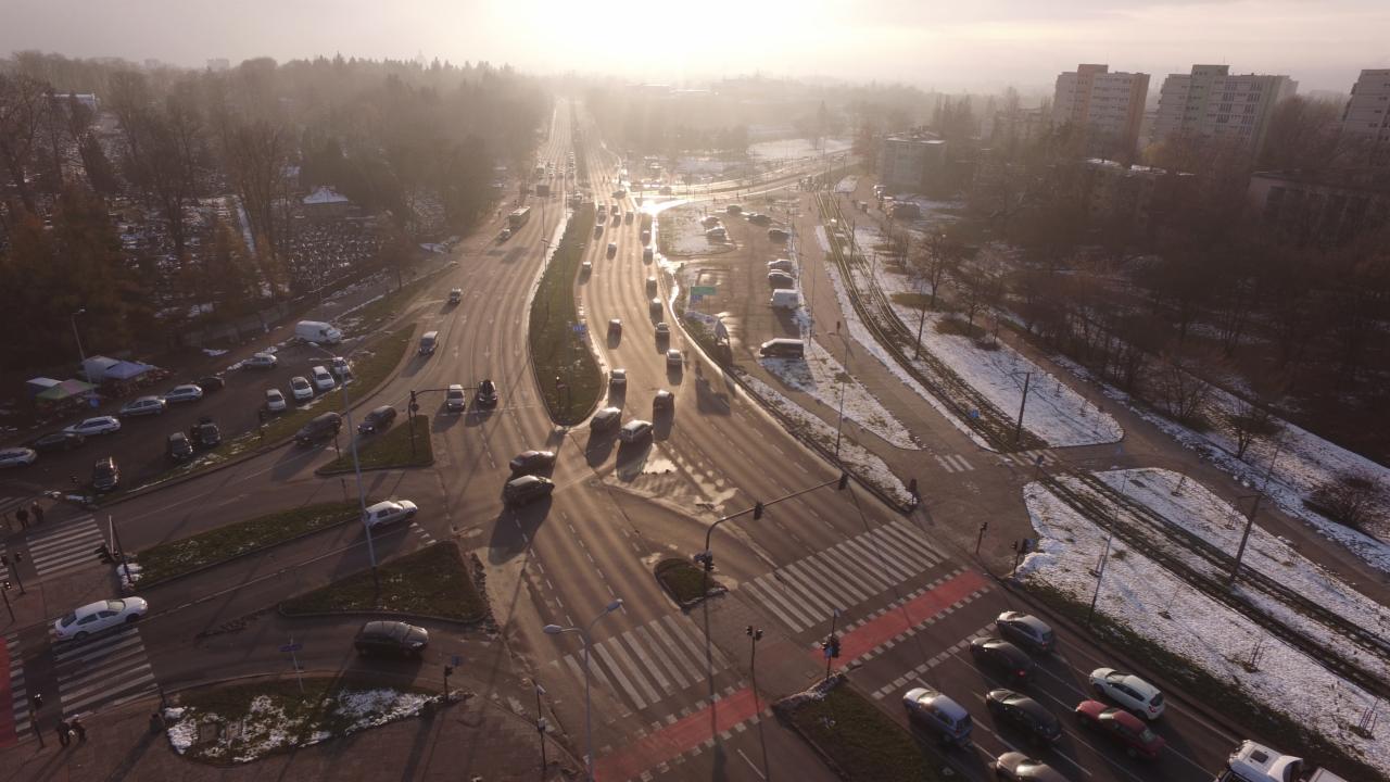 Zdjęcie z drona przedstawia skrzyżowanie ulic w mieście, z widocznymi samochodami, przejściami dla pieszych, torami tramwajowymi i budynkami w tle, wszystko w zimowej scenerii z delikatną mgłą.