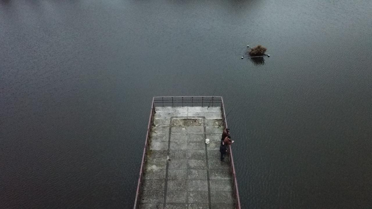 Widok z góry na betonowy pomost z dwoma wędkarzami łowiącymi ryby w pochmurny dzień, z widoczną roślinnością na wodzie.