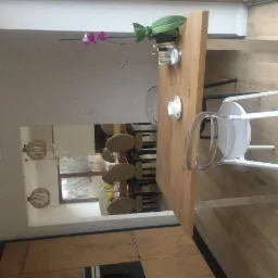 Jasne wnętrze jadalni z drewnianym stołem, białymi krzesłami z transparentnym oparciem, orchideą w wazonie i widokiem na salon w lustrze.