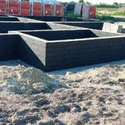 Czarne bloczki fundamentowe tworzące ściany budynku w trakcie budowy, widoczne pręty zbrojeniowe, ziemia i palety z cegłami w tle.