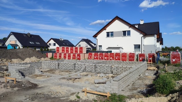 Budowa domu murowanego: wznoszenie fundamentów z bloczków betonowych, palety z czerwonymi cegłami w tle, widoczny fragment nowego osiedla.