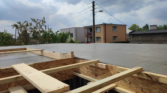 Świeżo wylany beton na stropie z widocznym szalunkiem drewnianym i budynkami w tle.