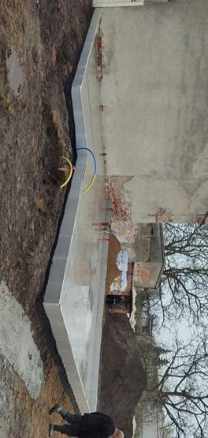 Świeżo wylany fundament z bloczków betonowych, widoczne wystające rury instalacyjne, tło stanowi ściana starego budynku z odsłoniętą cegłą.