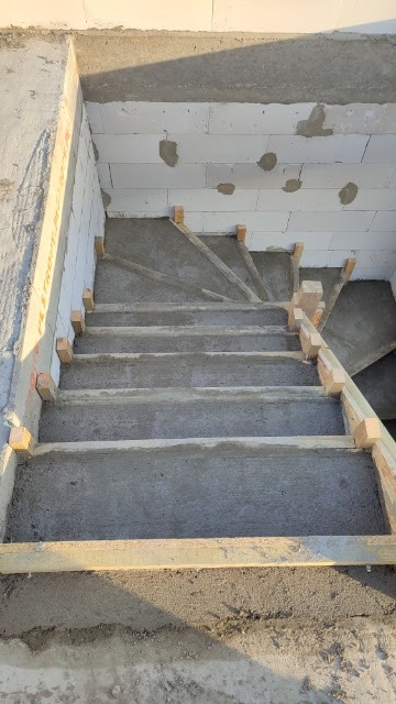 Wewnętrzne schody w surowym stanie, z drewnianym szalunkiem i częściowo otynkowanymi ścianami z białych bloczków, w trakcie budowy.