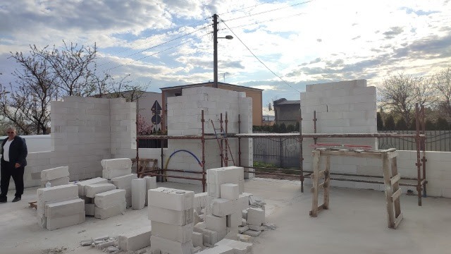 Wznoszenie ścian domu z bloczków betonu komórkowego, widoczne rusztowania i stosy materiału, mężczyzna w garniturze obserwuje postęp prac.