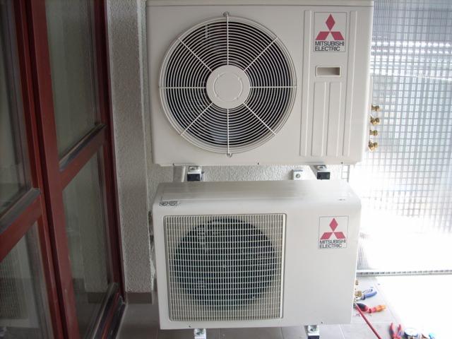 Dwa białe urządzenia klimatyzacyjne marki Mitsubishi Electric zamontowane jeden nad drugim na balkonie, widoczne narzędzia serwisowe obok dolnego urządzenia.