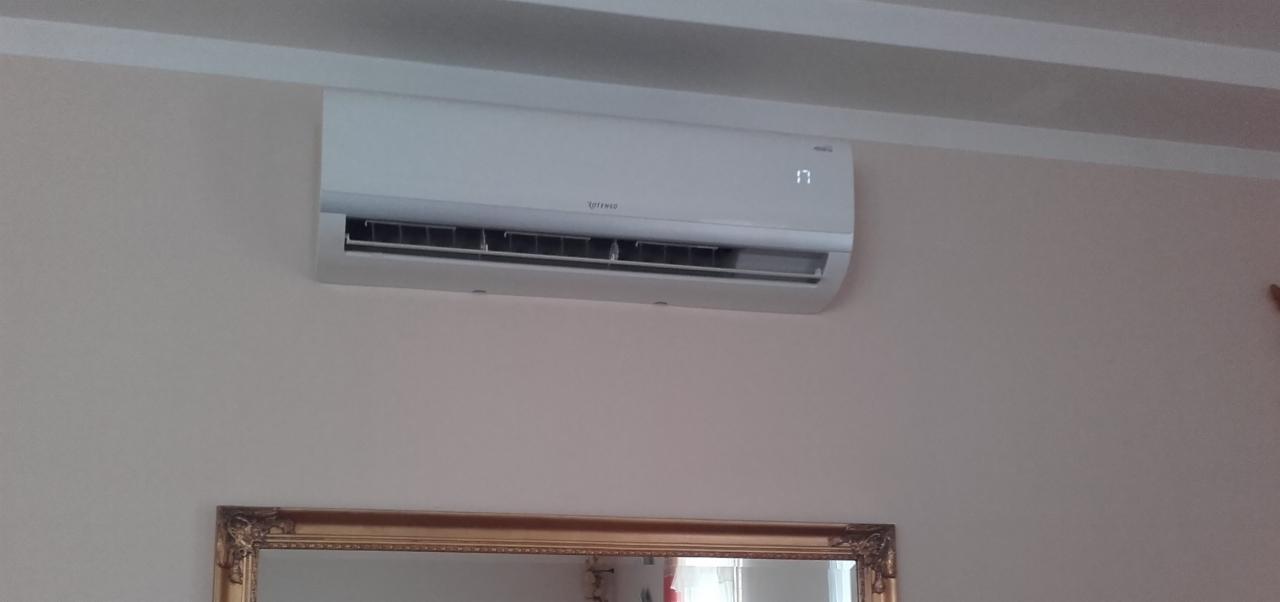 Biały klimatyzator Rotenso z wyświetlaczem temperatury 17 stopni, zamontowany na ścianie nad lustrem w złotej ramie.