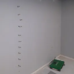 Świeżo osuszona ściana z widocznymi śladami po kołkach, przygotowana do dalszych prac wykończeniowych; w rogu pomieszczenia zielona skrzynka narzędziowa.