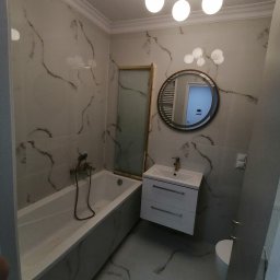 Adam Słodziński - Elegancka łazienka po remoncie: białe marmurowe płytki, wanna z zabudową, okrągłe lustro i złota armatura. Nowoczesne oświetlenie sufitowe.