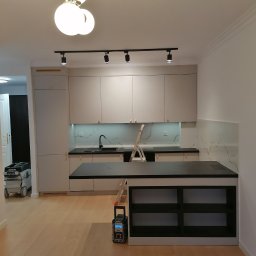 Adam Słodziński - Jasna kuchnia z wyspą i marmurowym backsplashiem. Widoczne narzędzia i drabina sugerują prace wykończeniowe. Podłoga z paneli, nowoczesne oświetlenie.