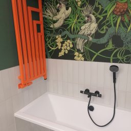 Adam Słodziński - Biała wanna z czarną armaturą i pomarańczowy grzejnik na tle ściany w tropikalne wzory. Nowoczesna aranżacja łazienki.
