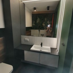 Adam Słodziński - Nowoczesna łazienka w Jeżowem z podświetlanym lustrem, umywalką nablatową i toaletą. W tle widoczna ściana z motywem roślinnym.