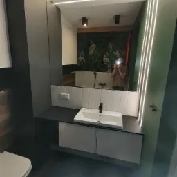 Nowoczesna łazienka w Jeżowem z podświetlanym lustrem, umywalką nablatową i toaletą. W tle widoczna ściana z motywem roślinnym.