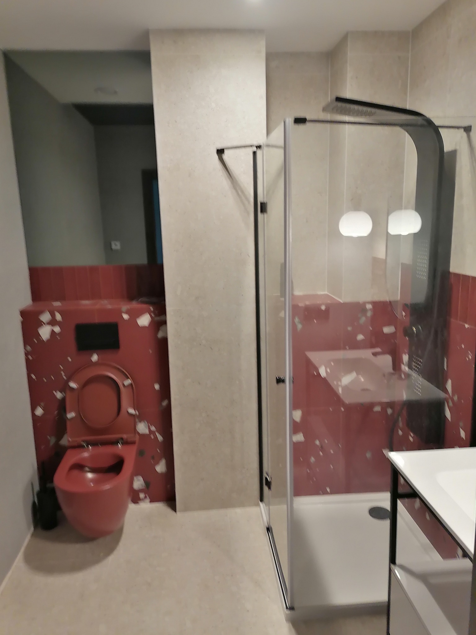 Nowoczesna łazienka po remoncie: czerwona armatura z motywem lastryko, kabina prysznicowa ze szklanymi drzwiami i biała umywalka. Nietypowe wykończenie.