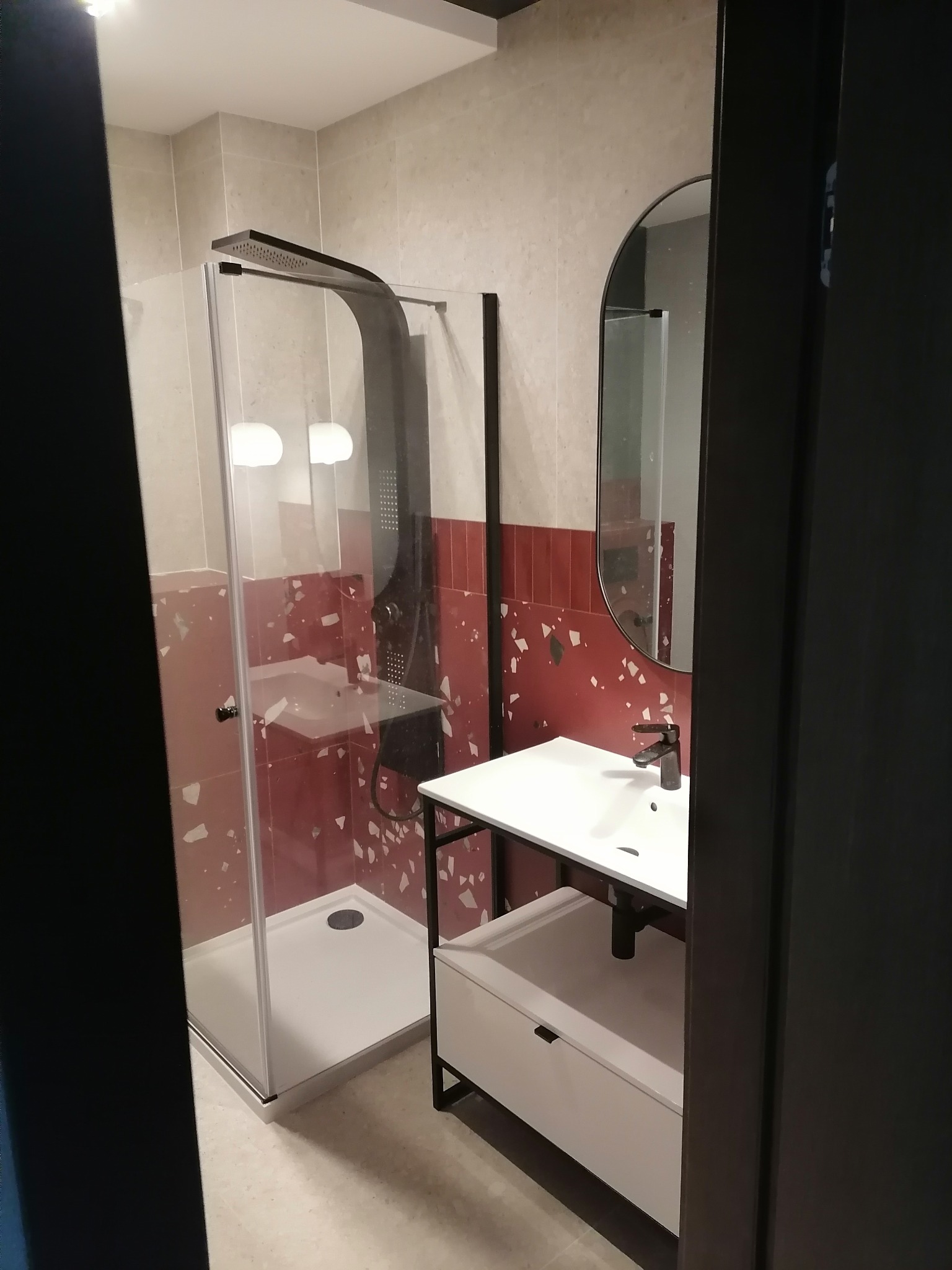 Nowoczesna łazienka z kabiną prysznicową typu walk-in i umywalką na metalowym stelażu. Czerwone płytki z białymi wzorami i duże lustro owalne w czarnej ramie.