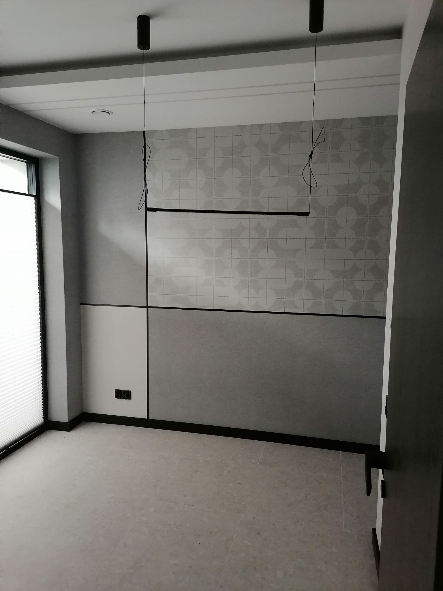 Minimalistyczne wnętrze: szara ściana z geometrycznym wzorem, nowoczesne lampy wiszące, jasna podłoga i okno z żaluzjami. Drzwi wejściowe po prawej stronie.