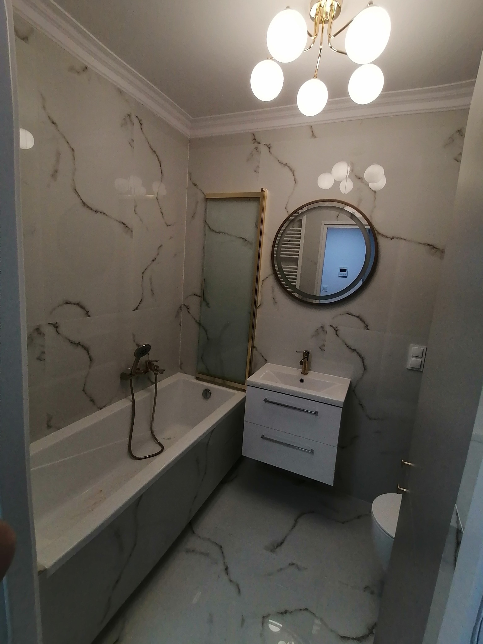 Elegancka łazienka po remoncie: białe marmurowe płytki, wanna z zabudową, okrągłe lustro i złota armatura. Nowoczesne oświetlenie sufitowe.