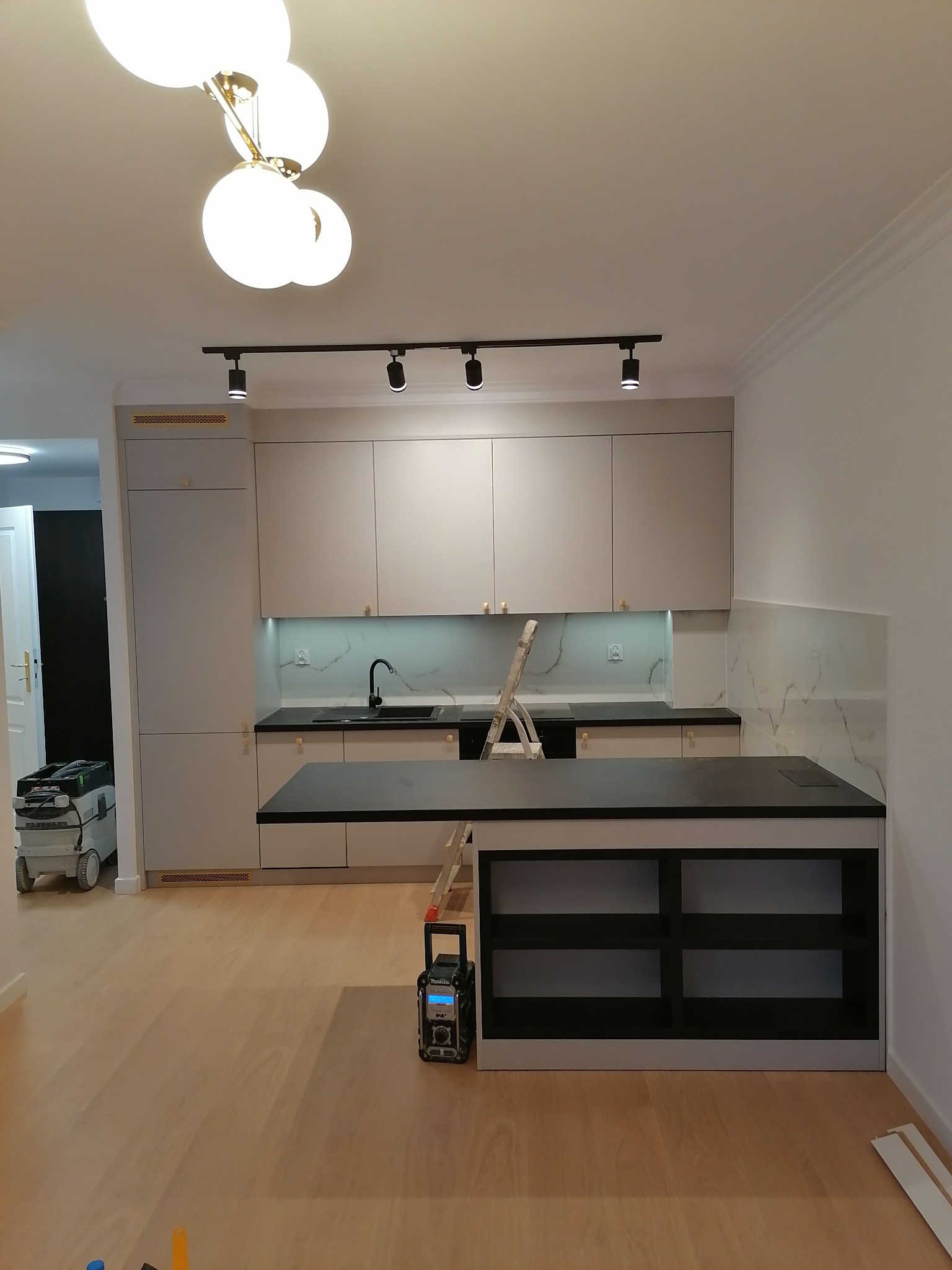 Jasna kuchnia z wyspą i marmurowym backsplashiem. Widoczne narzędzia i drabina sugerują prace wykończeniowe. Podłoga z paneli, nowoczesne oświetlenie.