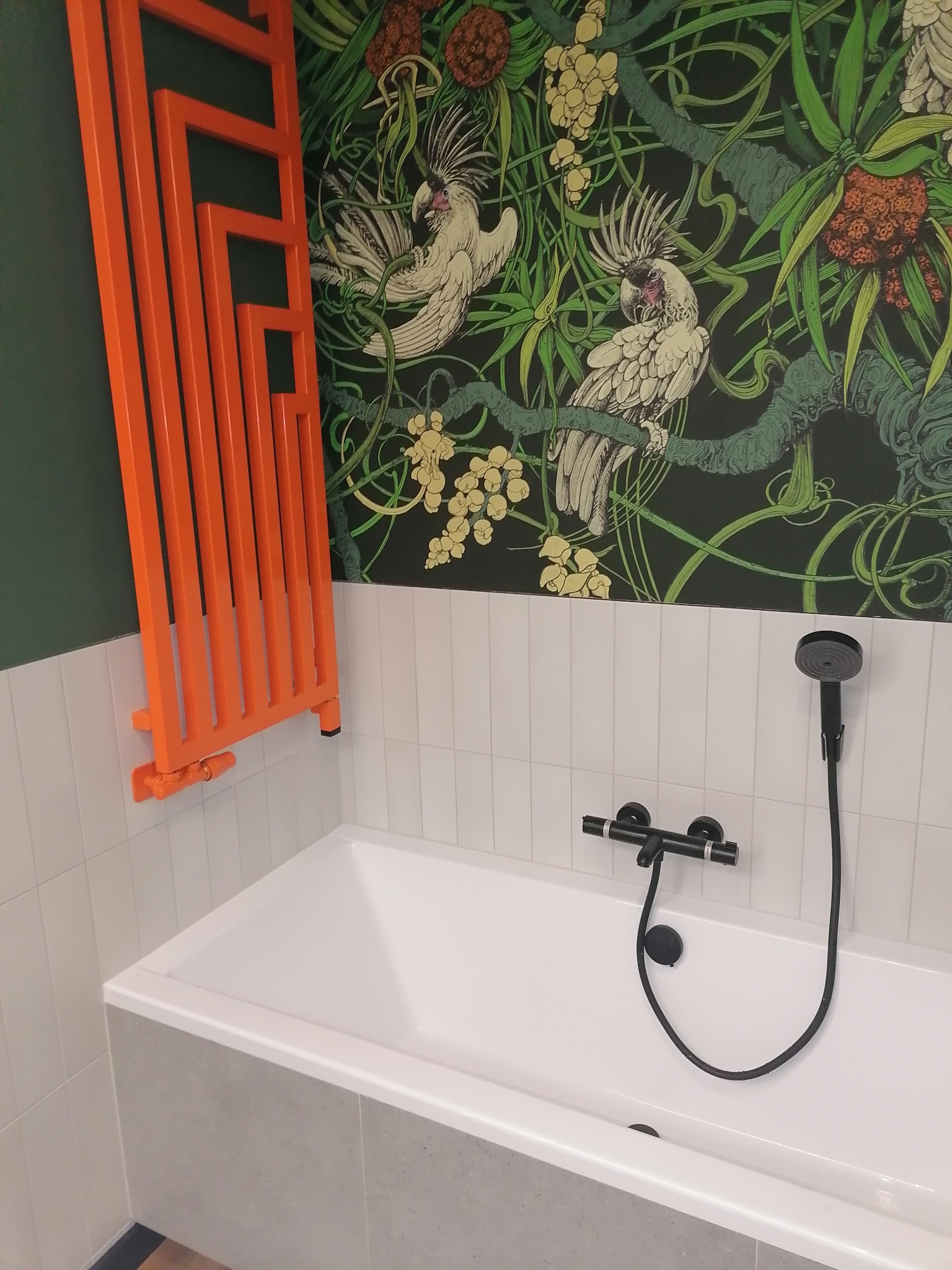 Biała wanna z czarną armaturą i pomarańczowy grzejnik na tle ściany w tropikalne wzory. Nowoczesna aranżacja łazienki.
