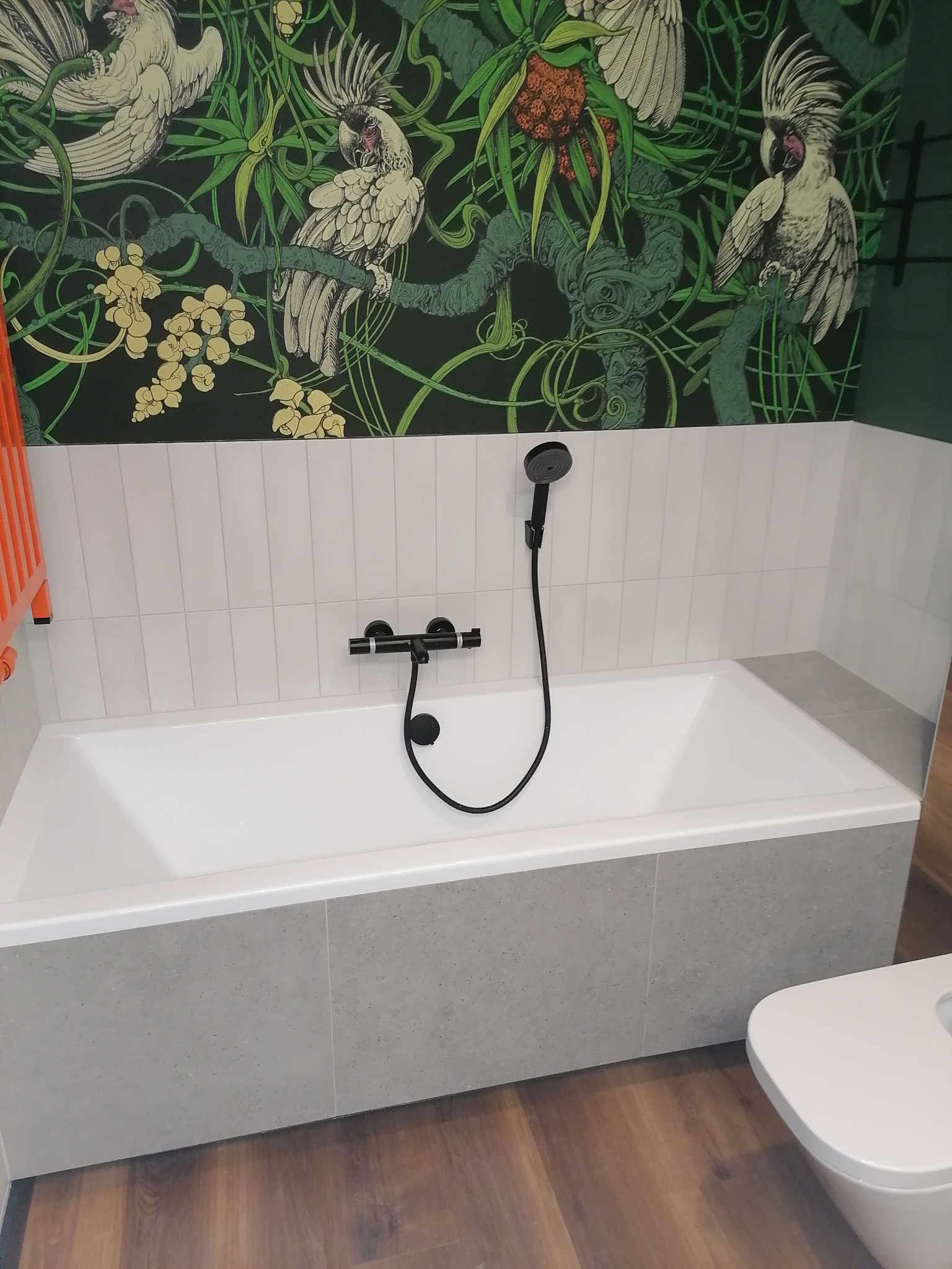 Nowoczesna łazienka z wanną obudowaną szarą płytką, białą armaturą i tapetą w tropikalne ptaki. Czarna bateria wannowa i prysznic.