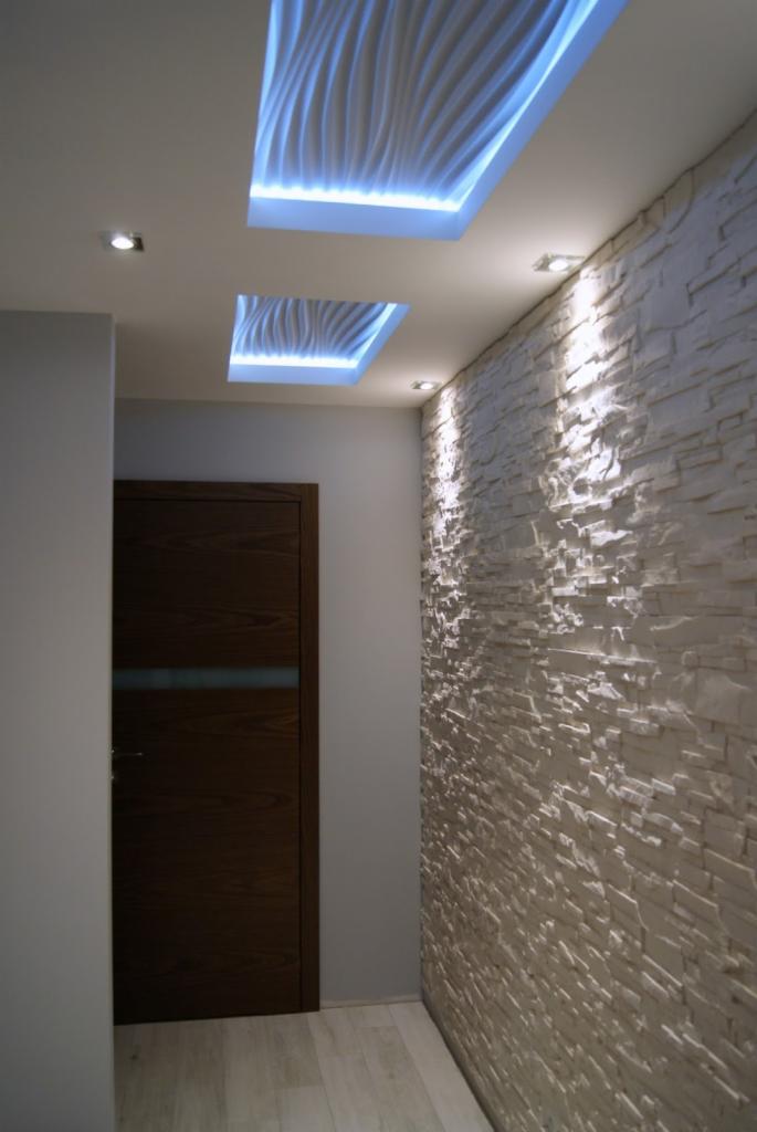 Nowoczesny przedpokój w Toruniu z dekoracyjną ścianą z białego kamienia, podświetlanymi panelami sufitowymi z motywem fal i drewnianymi drzwiami z poziomym przeszkleniem.