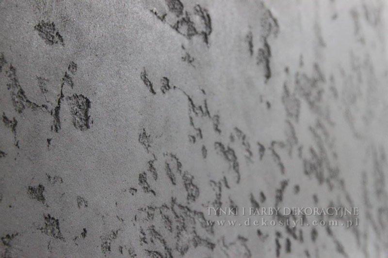 Szara ściana pokryta dekoracyjnym tynkiem o nieregularnej fakturze z widocznymi wżerami i nierównościami, ujęcie pod kątem, z widocznym adresem strony internetowej producenta tynków.