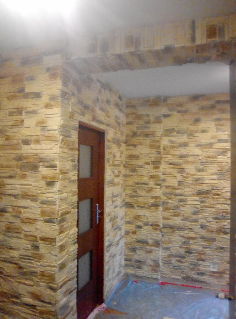 Wnętrze wykończone panelami ściennymi imitującymi kamień, z widoczną drewnianą ościeżnicą drzwiową i zabezpieczoną folią podłogą.