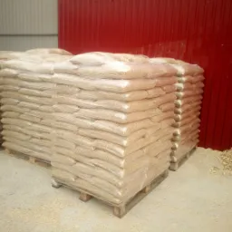 Stosy worków z pelletem, zabezpieczone folią, ułożone na paletach w magazynie z czerwoną ścianą. Obok widoczne zrębki drzewne.