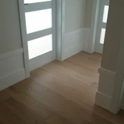 Jasne panele podłogowe ułożone w korytarzu z białymi ścianami i drzwiami z matowego szkła.