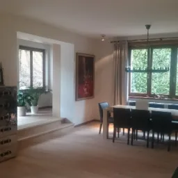 Jasne wnętrze domu z drewnianą podłogą, widok na jadalnię z oknem na ogród, dekoracyjny kominek z cegły i schodki do niższego poziomu z roślinami doniczkowymi.