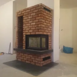 Murowany kominek z cegły klinkierowej z czarnym wkładem kominkowym i blatem, w surowym wnętrzu z widocznymi narzędziami i wiadrami po farbie.