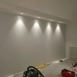 Wnętrze po szpachlowaniu i malowaniu na szaro, widoczne narzędzia malarskie, grzejnik i oświetlenie sufitowe