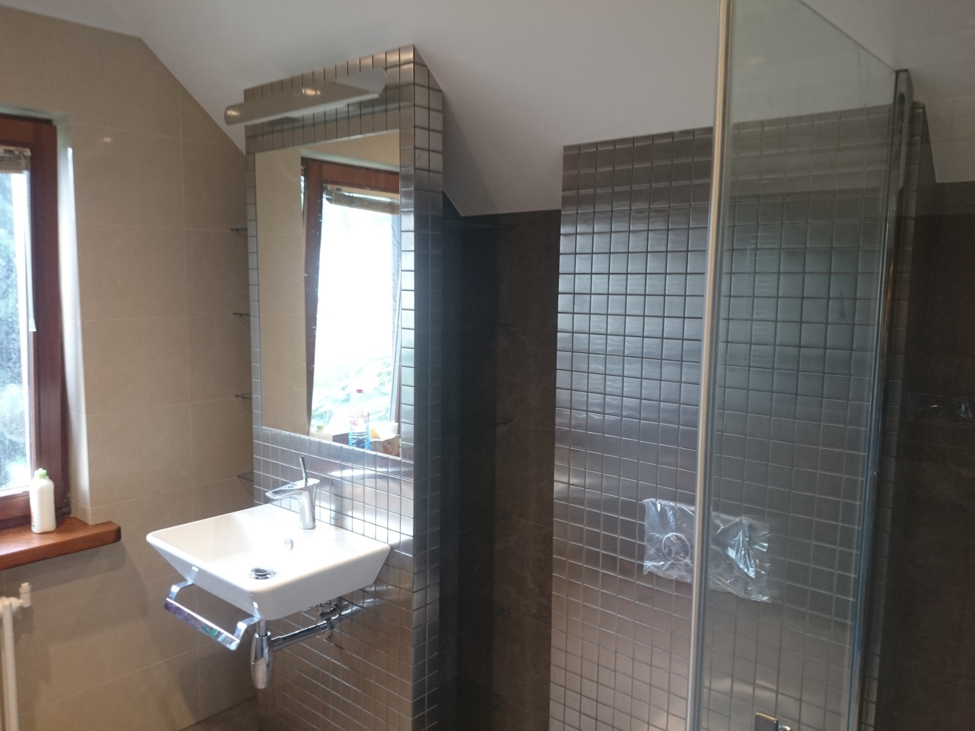 Ukończona łazienka w odcieniach szarości z mozaiką, widoczna umywalka, lustro z oświetleniem i kabina prysznicowa z folią ochronną na armaturze.