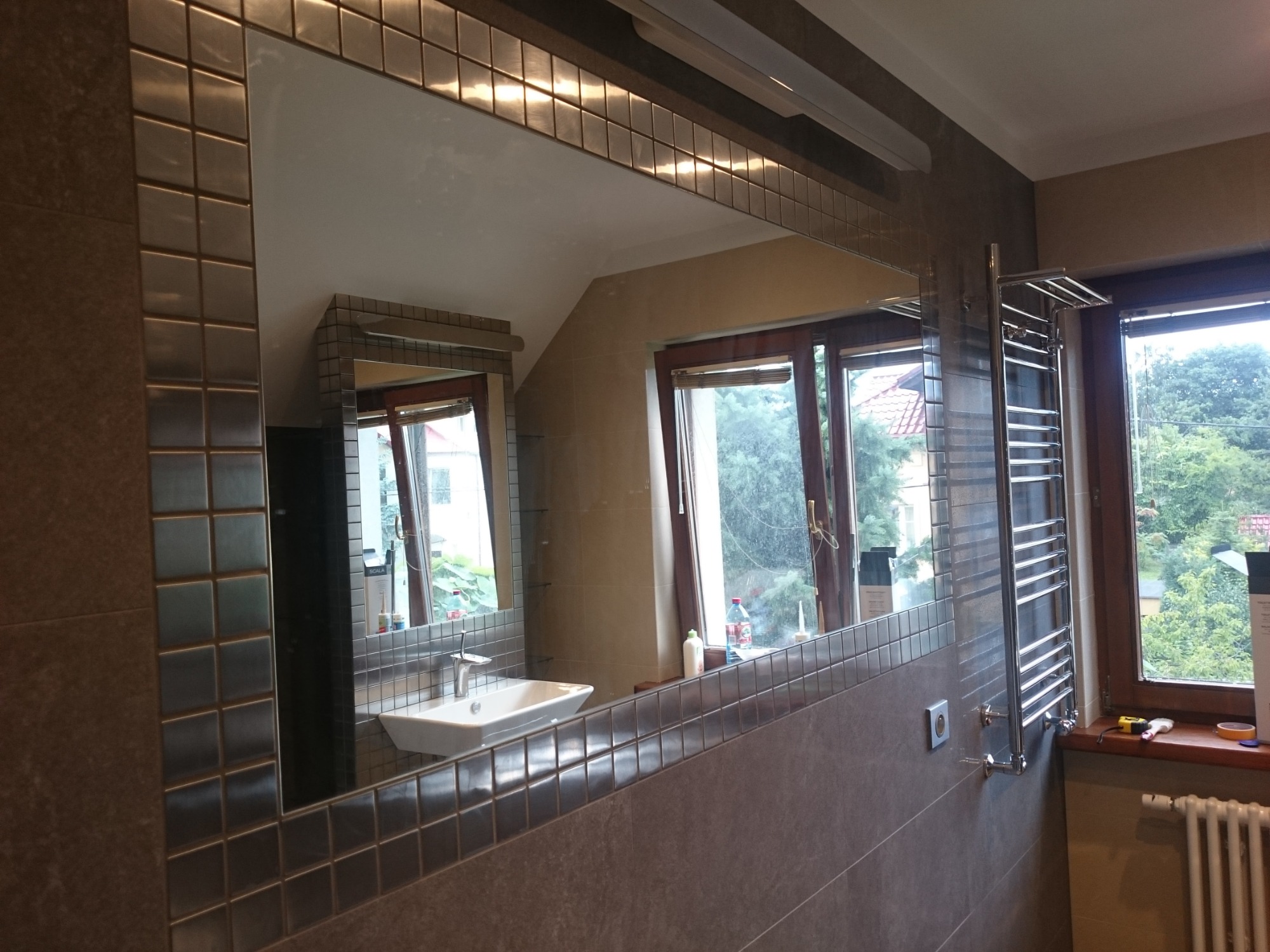 Nowoczesna łazienka z lustrem obłożonym mozaiką, grzejnikiem drabinkowym i dużym oknem z widokiem na zieleń.