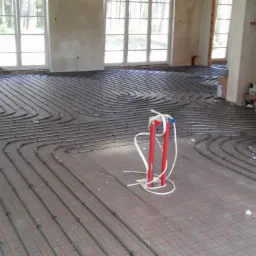 Montaż rur ogrzewania podłogowego w surowym wnętrzu, widoczne rury ułożone na siatce, rozdzielacz i materiały budowlane w tle.