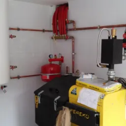 Biały bojler wiszący na ścianie z jasnych płytek, obok czerwony zbiornik wyrównawczy i żółto-czarny piec Pereko, widoczne rury miedziane i elementy instalacji grzewczej.