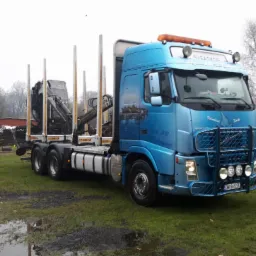 Niebieska ciężarówka Volvo z dźwigiem do transportu drewna na podmokłym terenie, w tle sterta drewna i budynki gospodarcze.