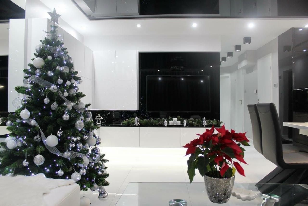 Nowoczesny salon w monochromatycznej bieli i czerni z akcentem świątecznym: choinka z srebrnymi ozdobami i poinsecja w błyszczącej doniczce.