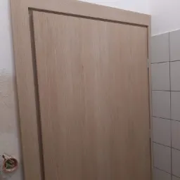 Zamknięte drzwi wewnętrzne z klamką i kluczem, widoczny fragment ściany z instalacją elektryczną i fragment ściany z białymi płytkami.