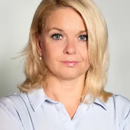 Portret kobiety z blond włosami, niebieskimi oczami i perłowym kolczykiem, ubrana w jasnoniebieską koszulę.