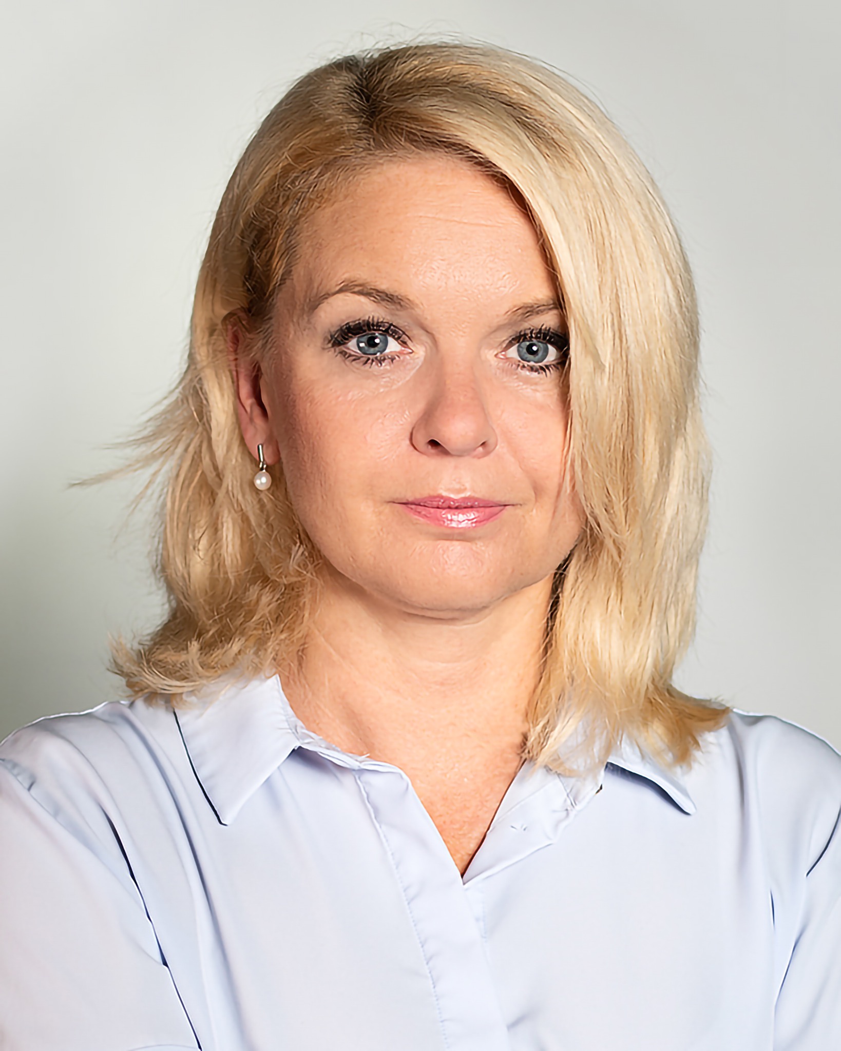 Portret kobiety z blond włosami, niebieskimi oczami i perłowym kolczykiem, ubrana w jasnoniebieską koszulę.