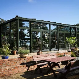 Ogród zimowy z ciemną ramą, widok z zewnątrz na drewniany stół i ławki, rośliny w donicach na ceglanym murku.