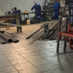 Warsztat obróbki metali: pracownicy w kombinezonach roboczych przy stanowiskach spawalniczych, porozrzucane profile metalowe na podłodze z płytek.