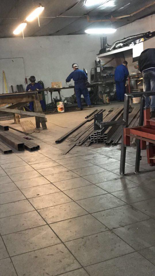 Warsztat obróbki metali: pracownicy w kombinezonach roboczych przy stanowiskach spawalniczych, porozrzucane profile metalowe na podłodze z płytek.