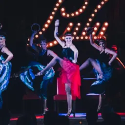 Zespół tancerek kankana w czarno-niebieskich kostiumach z piórami i cekinami, wykonujących synchroniczny układ na scenie z czerwonym oświetleniem i dekoracyjnym napisem 'Lady' z żarówek w tle.