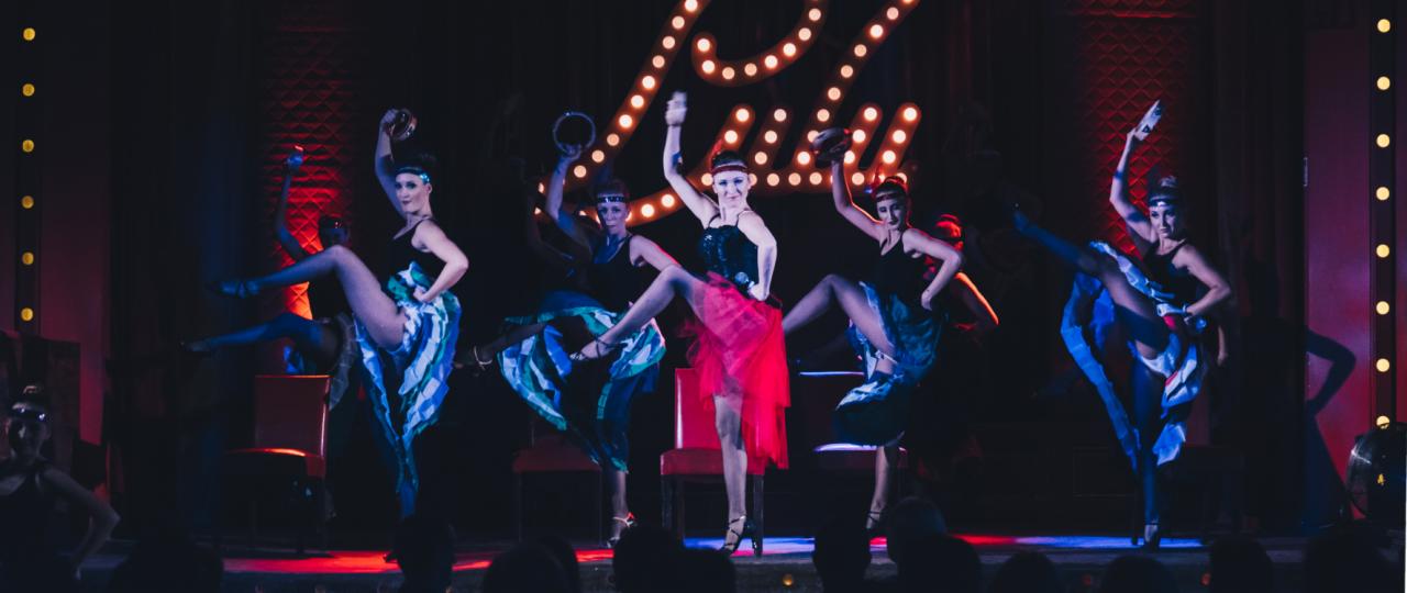 Zespół tancerek kankana w czarno-niebieskich kostiumach z piórami i cekinami, wykonujących synchroniczny układ na scenie z czerwonym oświetleniem i dekoracyjnym napisem 'Lady' z żarówek w tle.