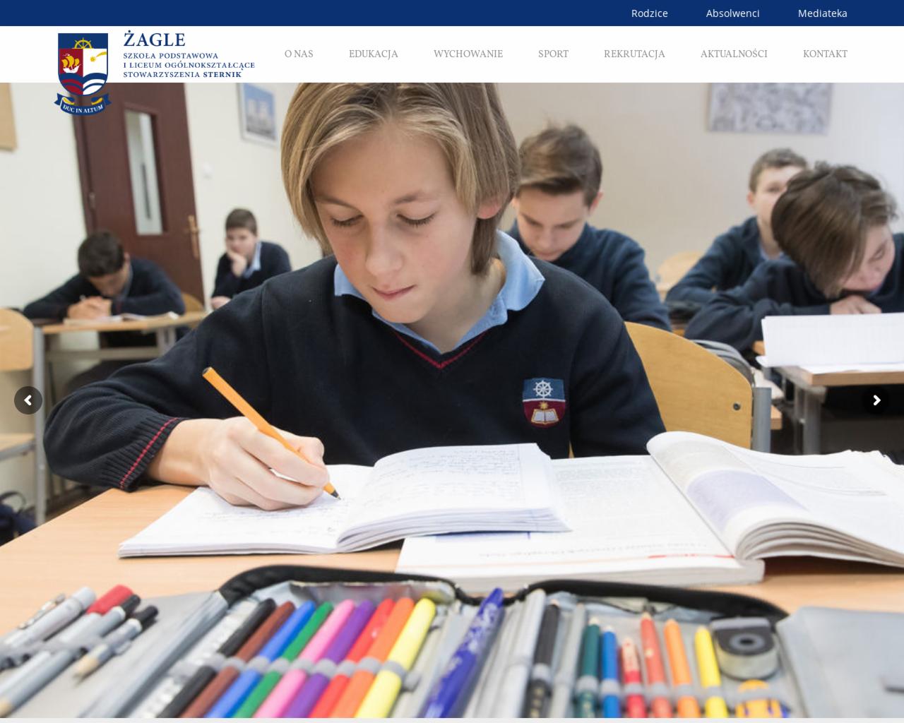 Uczeń pisze w zeszycie na tle innych uczniów w klasie. Na pierwszym planie otwarty piórnik z kolorowymi pisakami.