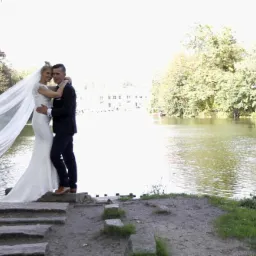 Para młoda w objęciach na tle jeziora i drzew, ona w białej sukni ślubnej z długim welonem, on w granatowym garniturze i brązowych butach, stoją na kamiennych stopniach.